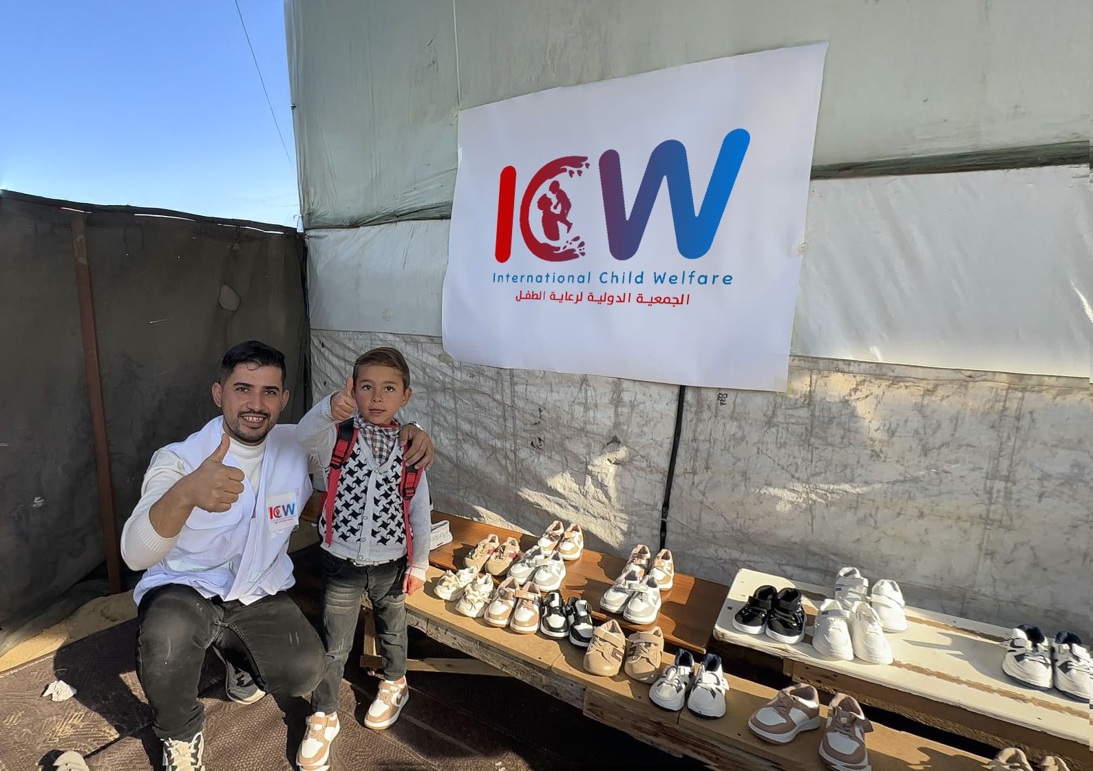 A humanitarian worker wearing a white vest with the "International Child Welfare" (ICW) logo gives a thumbs-up while posing with a young child who is also giving a thumbs-up. They are in an outdoor setting with a white tent in the background. A banner with the ICW logo hangs on the tent, and several pairs of children's shoes are displayed on wooden tables.