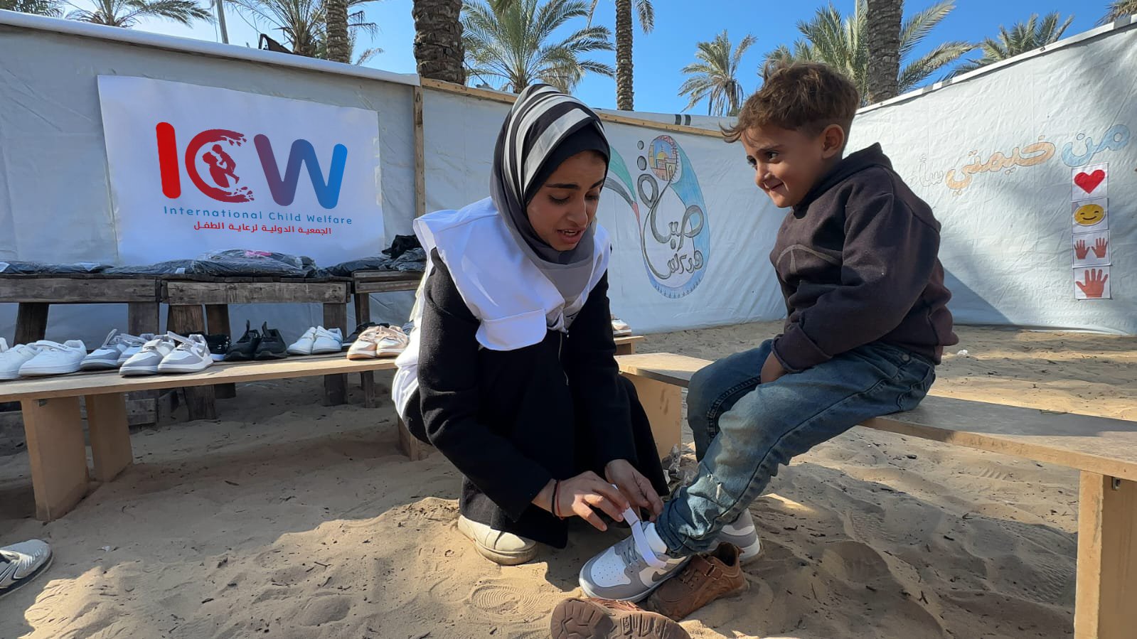 A young woman wearing a black outfit, a white vest, and a striped hijab is kneeling on sandy ground, helping a smiling young boy put on a new pair of sneakers. The boy, dressed in a dark sweater and jeans, is sitting on a wooden bench, looking happy. Behind them, there is a banner with the logo of 'International Child Welfare' in both English and Arabic, along with a display of shoes and clothing on wooden tables. The setting appears to be an outdoor humanitarian aid distribution event, with palm trees and a bright blue sky in the background. Another banner with Arabic text and images of hands and hearts is visible on a tent behind them.